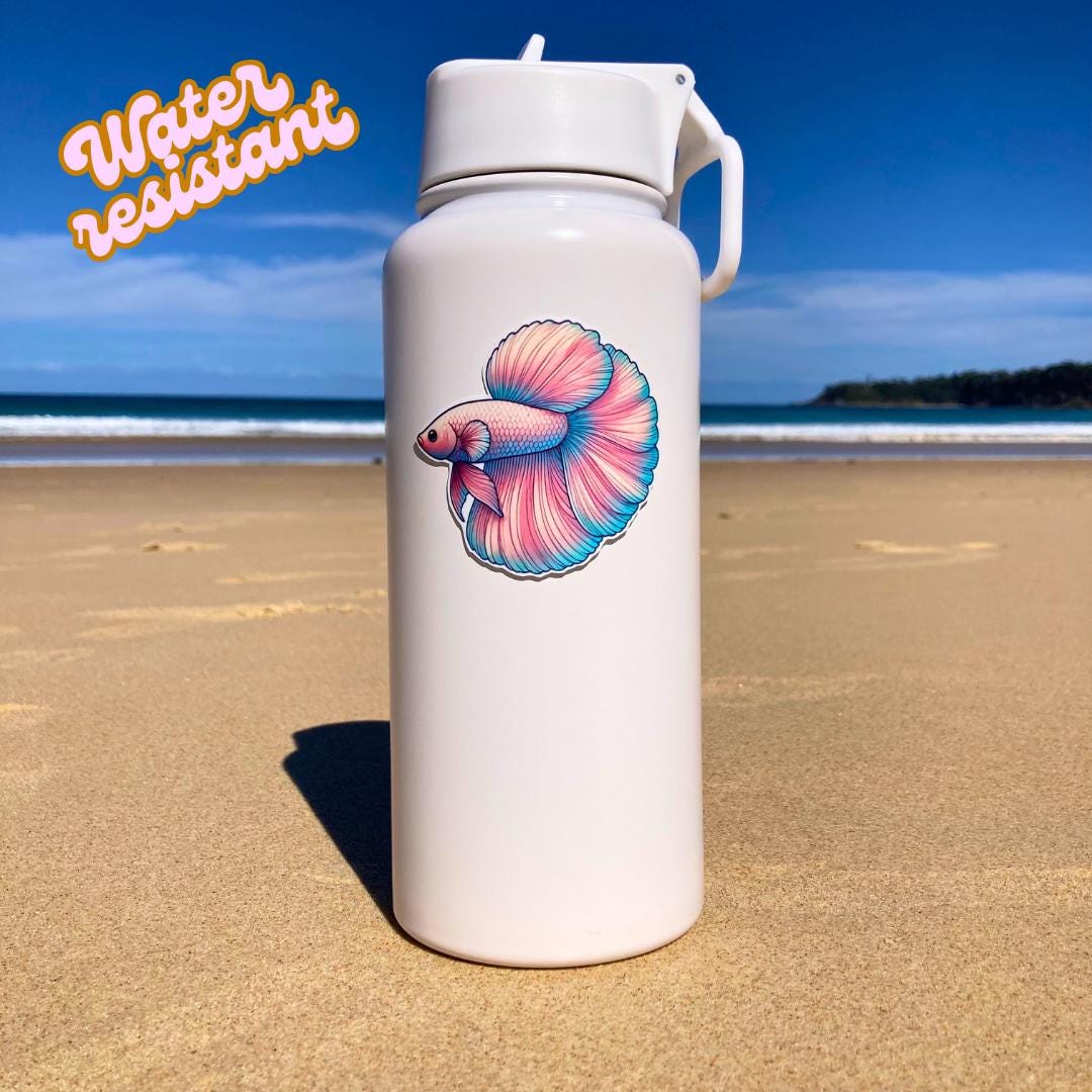A white water bottle with a colorful betta fish sticker is placed on a sandy beach with a blue ocean and clear sky in the background. The words "Water resistant" in orange and white are in the top left corner, emphasizing the sticker's durability.
