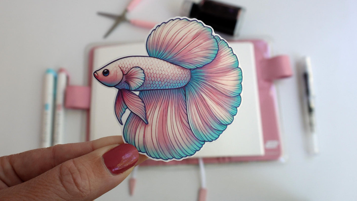 A hand holds a sticker of a betta fish with flowing fins and scales in soft shades of pink, teal, and purple. In the background, there is a pink journal, colored pens, and stationery, creating a neat and creative workspace setting.