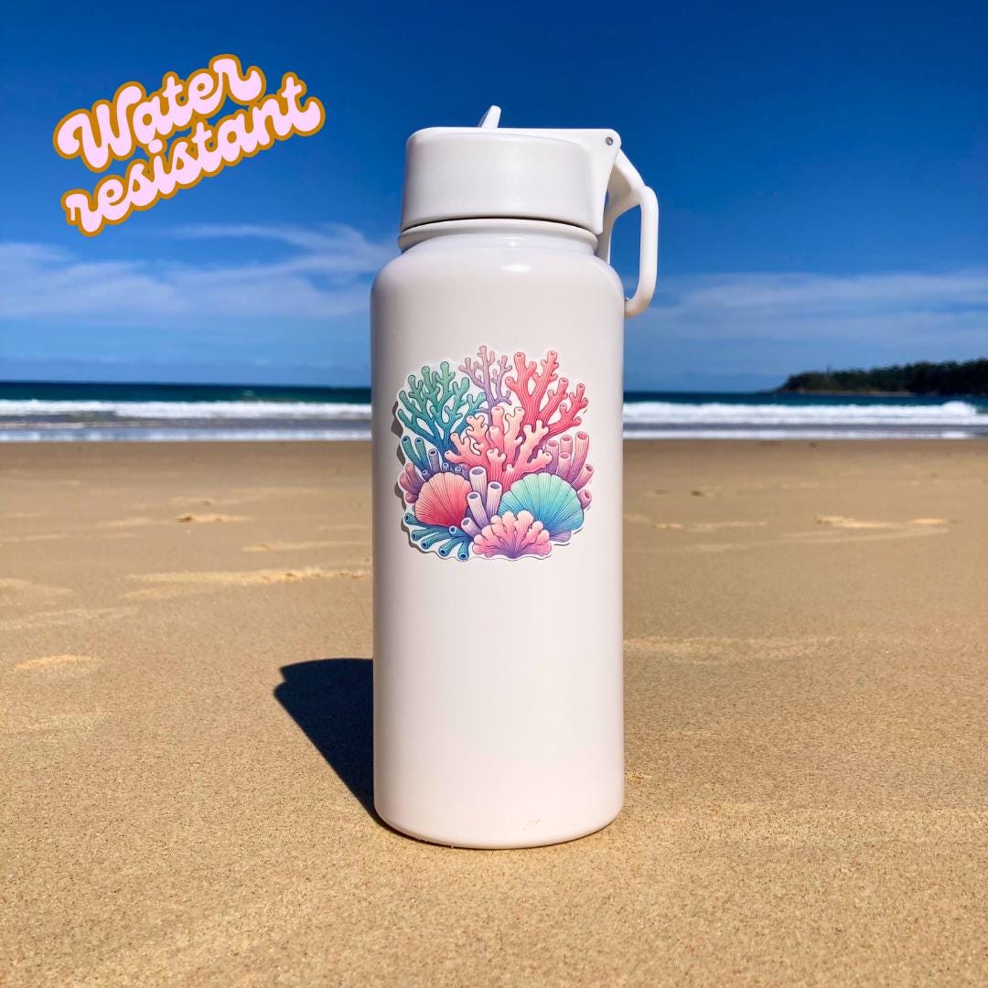 A white water bottle with a colorful coral reef sticker featuring pink, blue, purple, and green corals is placed on a sandy beach. The background shows the ocean and a clear sky. The words "Water resistant" in orange text are in the top left corner.