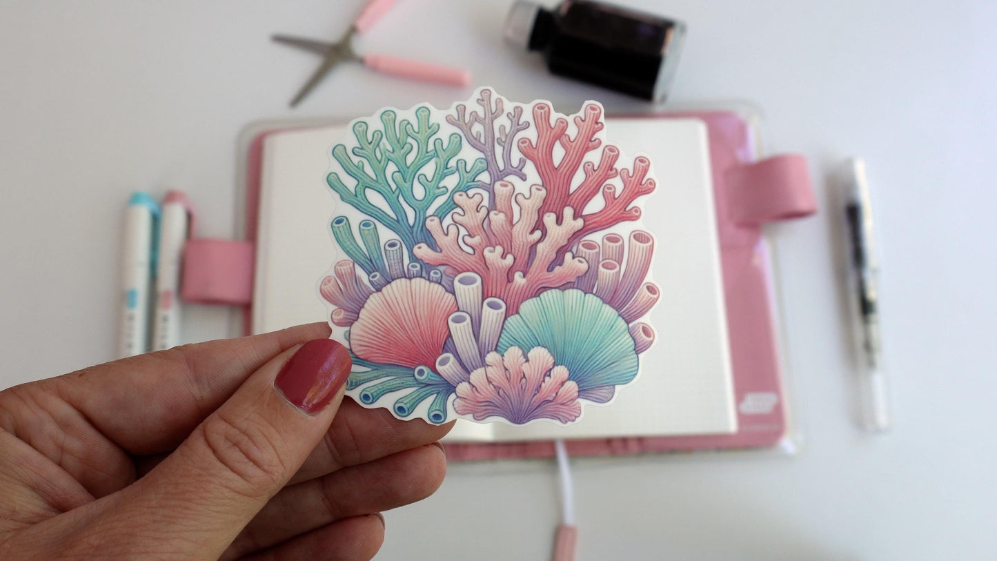 A hand holds a sticker of colorful coral and seashells in shades of pink, teal, and purple. In the background, a pink notebook, colored pens, and crafting tools are visible, creating a creative workspace setting.