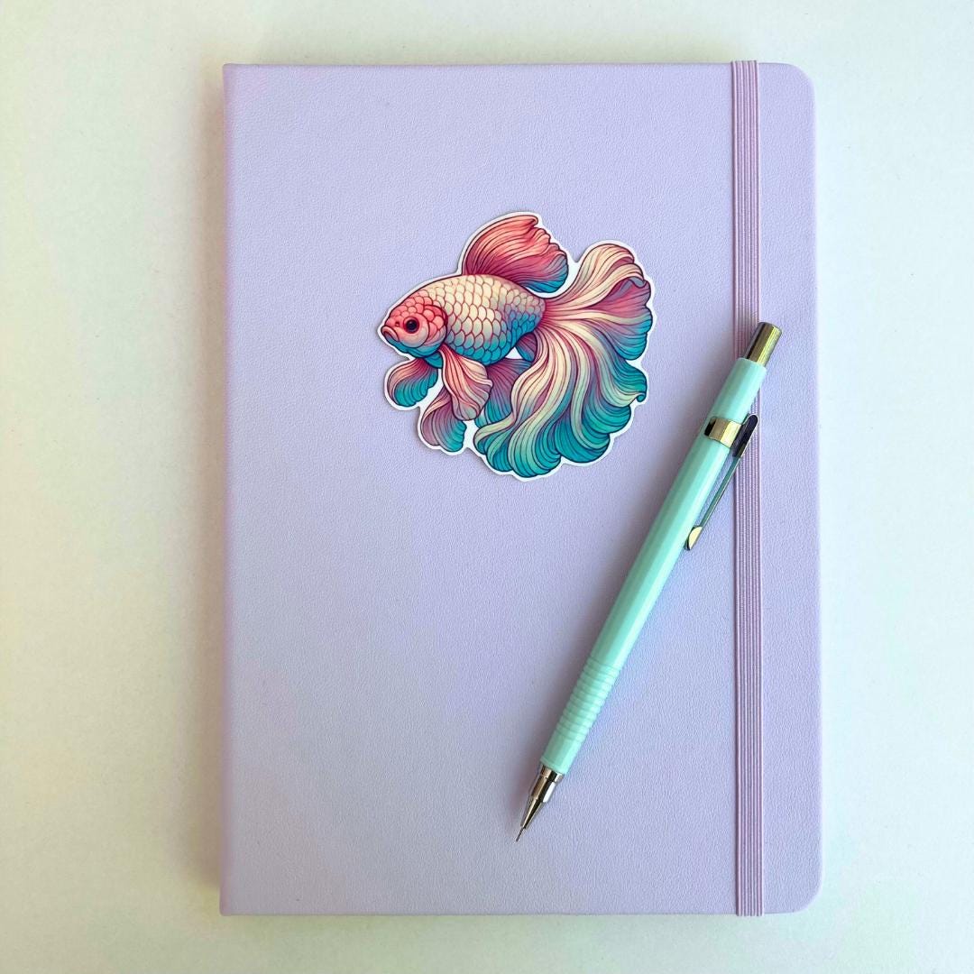 A pastel purple notebook with an elastic band features a colorful betta fish sticker in pink, blue, and green hues. A light blue mechanical pencil with gold accents rests diagonally on top. The simple background highlights the stationery items.