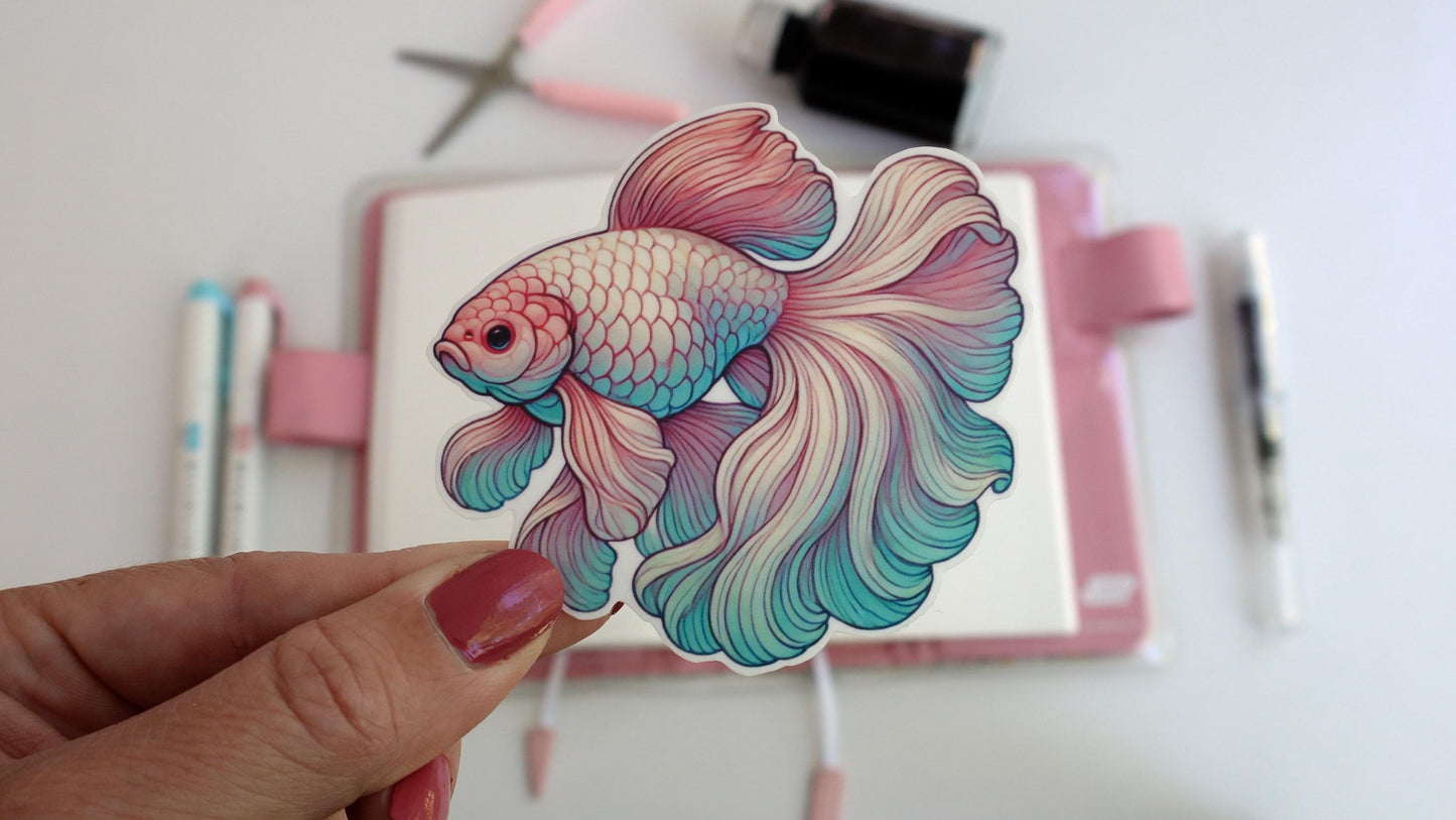 A hand holds a sticker of a colorful betta fish with flowing fins in shades of pink, teal, and purple. In the background, a pink notebook, pens, scissors, and ink bottle are visible, creating a workspace scene.