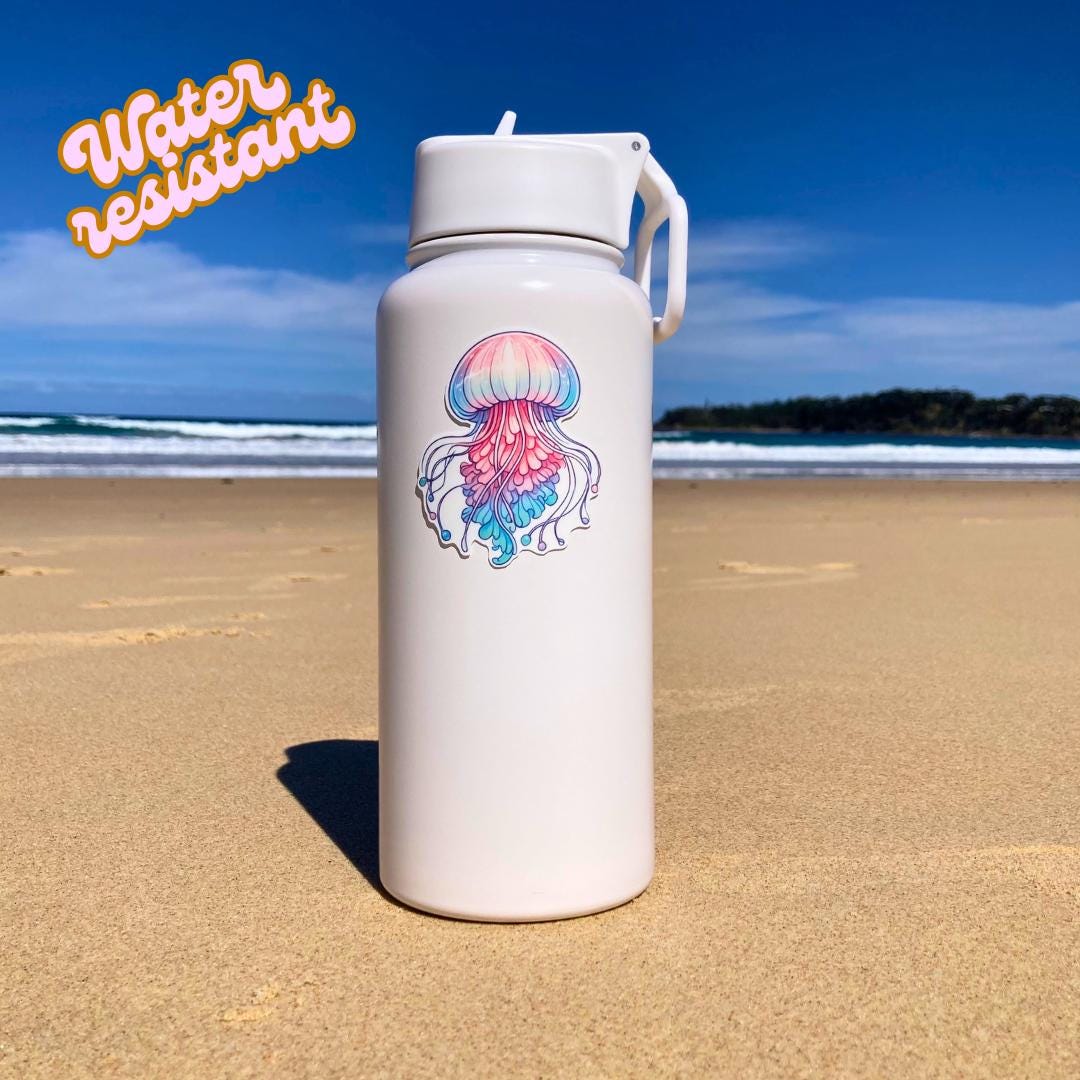 A white water bottle with a colorful jellyfish sticker in pink, blue, and purple hues is placed on a sandy beach. The background shows the ocean and a clear sky. The words "Water resistant" are in the top left corner, highlighting durability