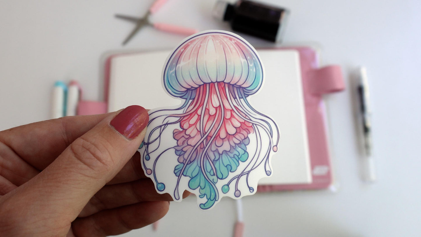 A hand holds a sticker of a pastel jellyfish with flowing tentacles in pink, purple, and teal. The background shows a desk setup with a pink notebook, pens, scissors, and an ink bottle, creating a creative workspace scene.
