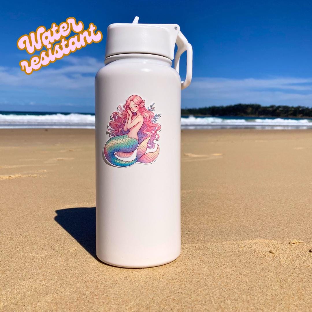 A white water bottle with a colorful mermaid sticker featuring pink hair and a blue-green tail is placed on a sandy beach. The background shows the ocean and a clear sky. The words "Water resistant" are in the top left corner, highlighting durability