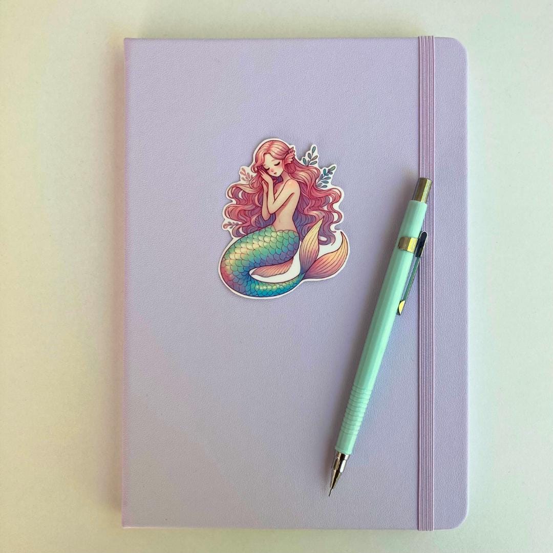 A pastel purple notebook with an elastic band features a mermaid sticker with pink hair and a blue green tail A light blue mechanical pencil with gold accents rests diagonally on top. The simple background highlights the notebook and stationery items