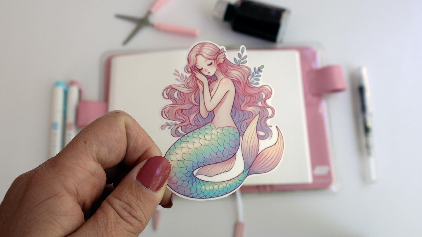 A hand holds a sticker of a serene pastel mermaid with long pink hair and a teal, lavender, and yellow scaled tail. The background shows a desk setup with pens, scissors, and an ink bottle, creating a creative workspace atmosphere.