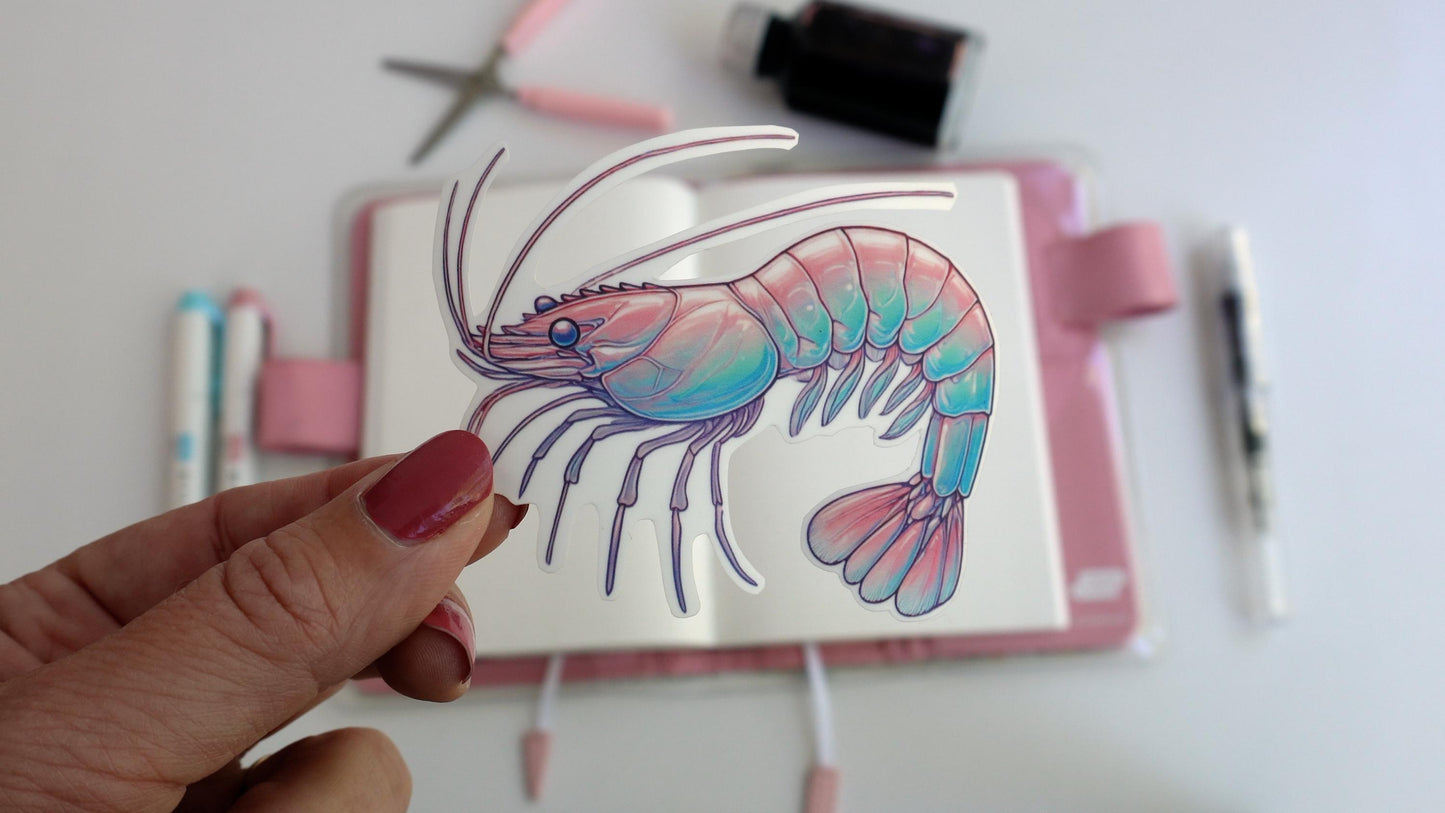 A hand holds a sticker of a pastel shrimp, featuring soft pink, blue, and purple hues. The background includes a notebook, pastel-colored pens, scissors, and an ink bottle, suggesting a creative workspace setup.