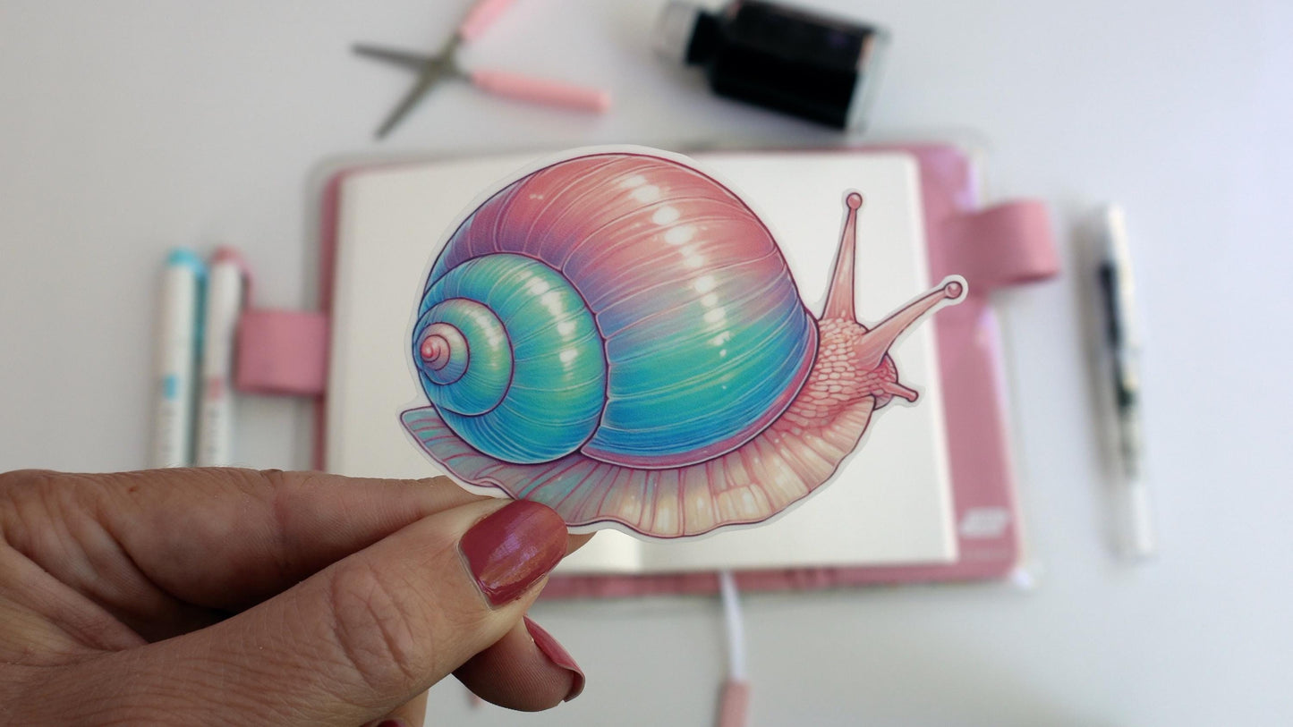 A hand holds a sticker of a pastel-colored snail with a pink, blue, and green gradient shell. In the background, a notebook, pastel-colored pens, scissors, and an ink bottle are arranged on a white desk, suggesting an art workspace.