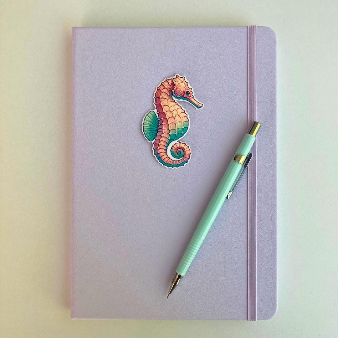 A pastel purple notebook with an elastic band features a colorful seahorse sticker in pink, blue, and green hues. A light blue mechanical pencil with gold accents rests diagonally on top. The minimal background highlights the notebook and pencil.