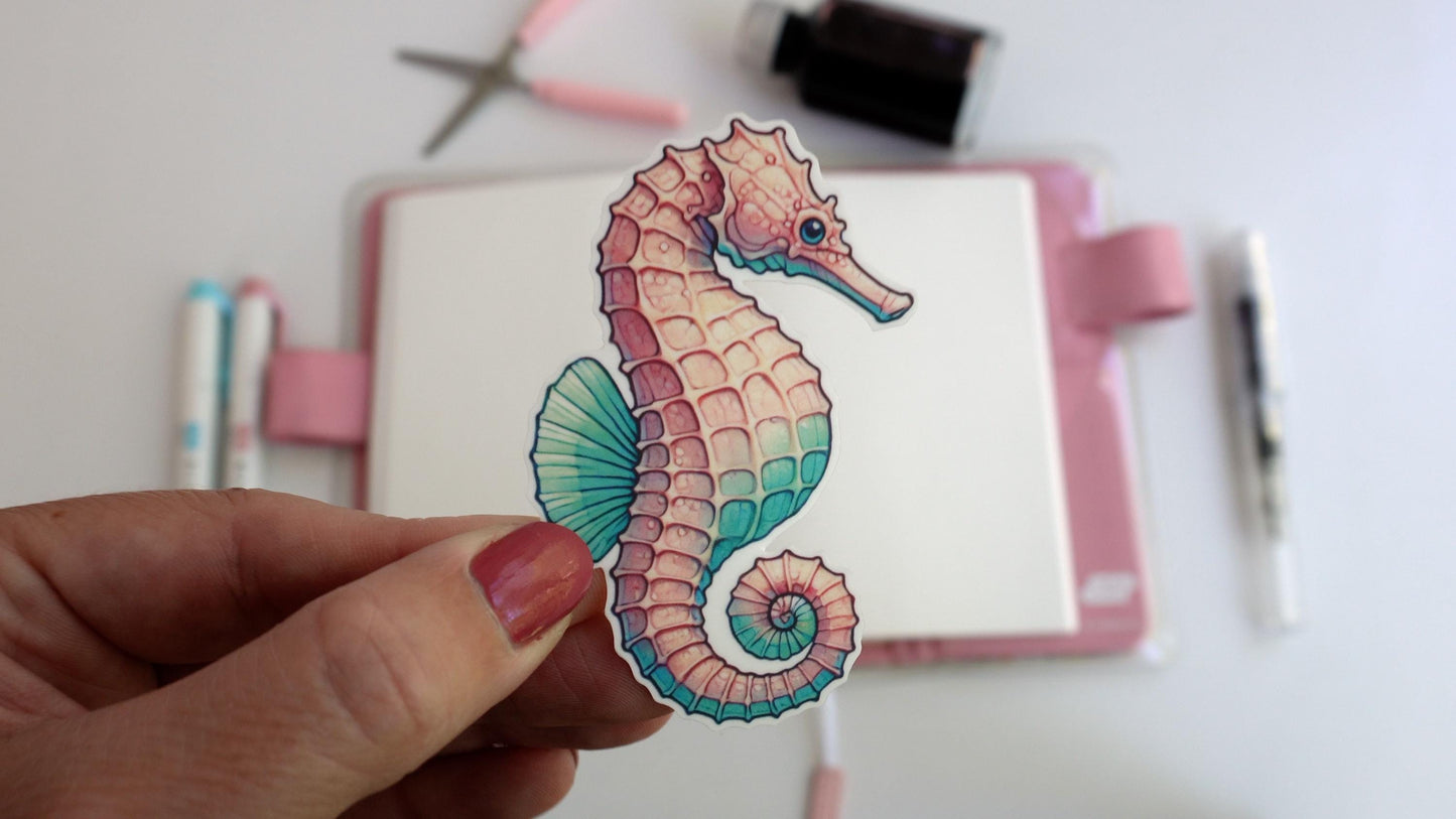 A hand holds a pink and teal seahorse sticker with intricate details. The background shows an artistic workspace with pens, scissors, and an ink bottle, alongside a closed notebook with a pink elastic band.