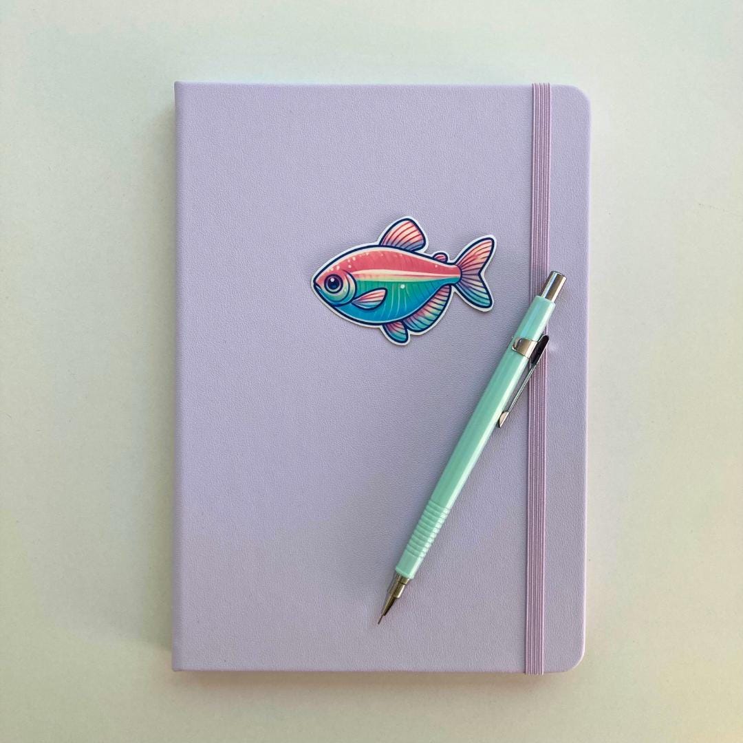 A pastel purple notebook is adorned with a colorful tetra fish sticker, featuring pink, teal, and blue hues. A mint green mechanical pencil with silver accents rests diagonally on the notebook’s right side, with a soft white background.