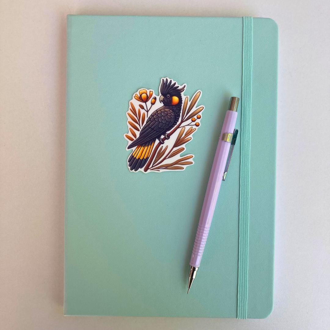 A light turquoise notebook with an elastic strap features a sticker of a black cockatoo perched on a branch with orange flowers and leaves. A lavender mechanical pencil is placed diagonally on the notebook, which rests on a white surface.