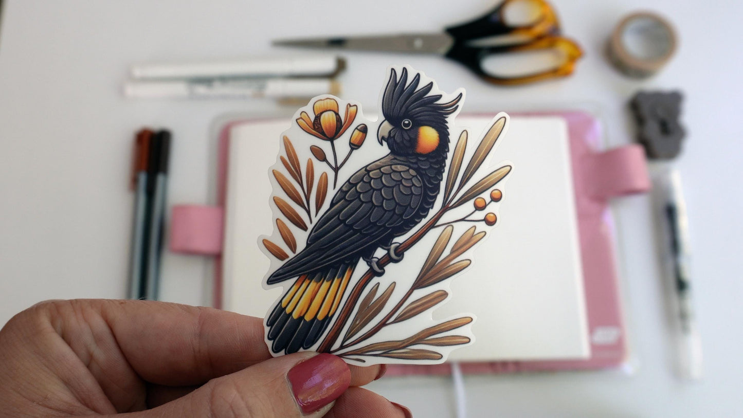 A hand holds a sticker of a black cockatoo perched on a branch with orange flowers and leaves. In the background, an open notebook with a pink cover, pens, scissors, and tape are visible.