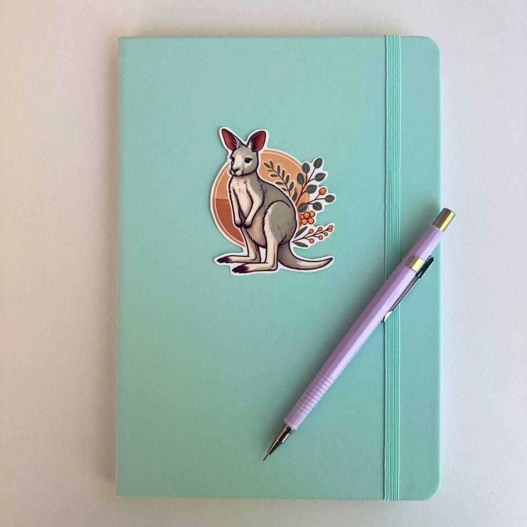 A light turquoise notebook with an elastic strap features a sticker of a cartoon kangaroo with a tan circle and leafy branches in the background. A lavender mechanical pencil is placed diagonally on the notebook, which lies on a white surface.