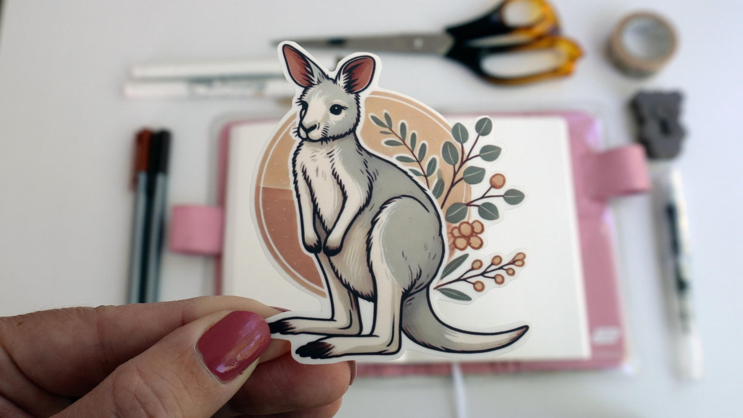 A hand holds a kangaroo sticker in front of an open notebook with a pink cover. The kangaroo is white with a tan circle and leafy branches behind it. The background includes scattered pens, scissors, and a roll of tape on a white surface