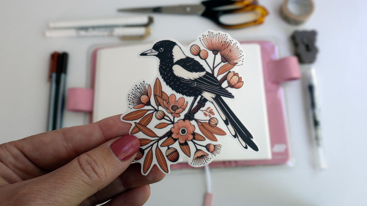A hand holds a sticker of a black and white magpie perched on a branch with orange flowers and leaves. Background includes scissors, pens, and stationery items, slightly blurred, with a pink notebook underneath.