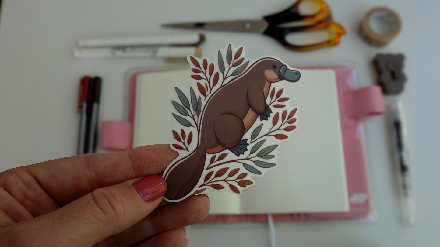 A hand holds a platypus sticker in front of an open notebook with a pink cover. Scattered on the white surface are pens, scissors, and a roll of tape, suggesting a creative workspace. The sticker features a platypus with leafy branches
