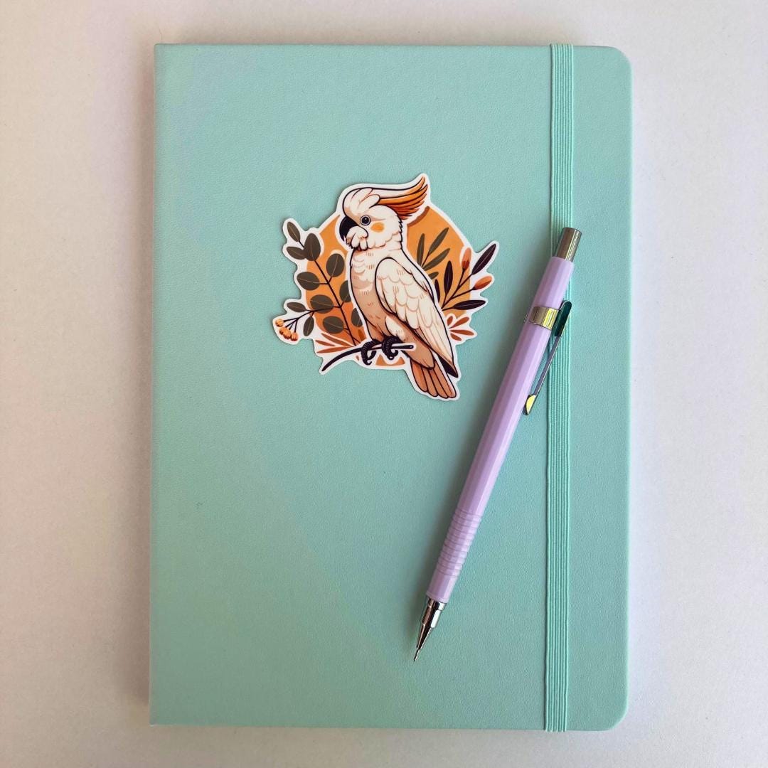 A pastel blue notebook with an elastic band is decorated with a sticker of a white cockatoo with an orange crest, perched on a branch surrounded by leaves. A lavender mechanical pencil with silver accents rests diagonally on the notebook's right side