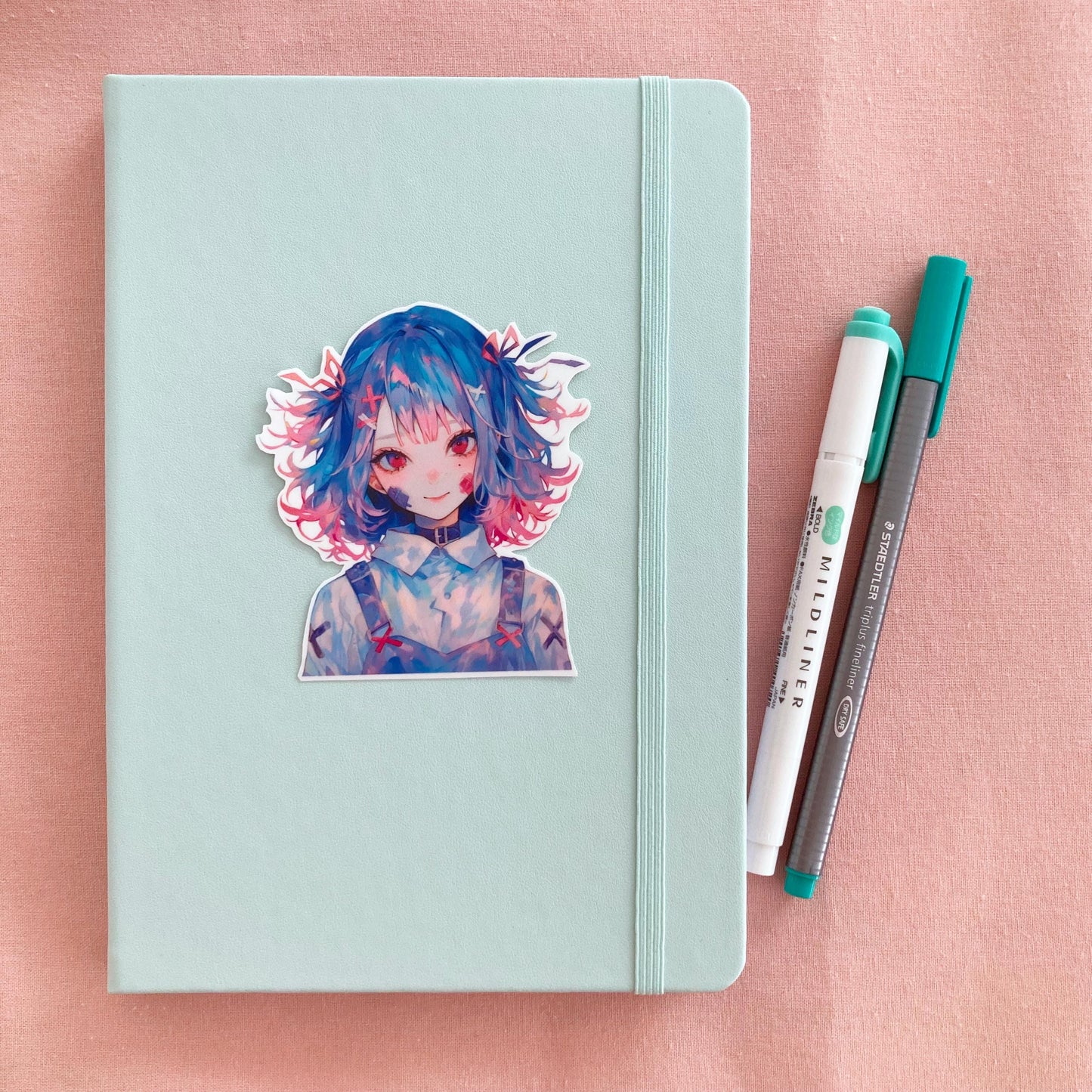 a blue and red haired anime girl sticker wearing overalls on the cover of a mint coloured journal next to a mint green mildliner pen and green felt tip pen on a pink background