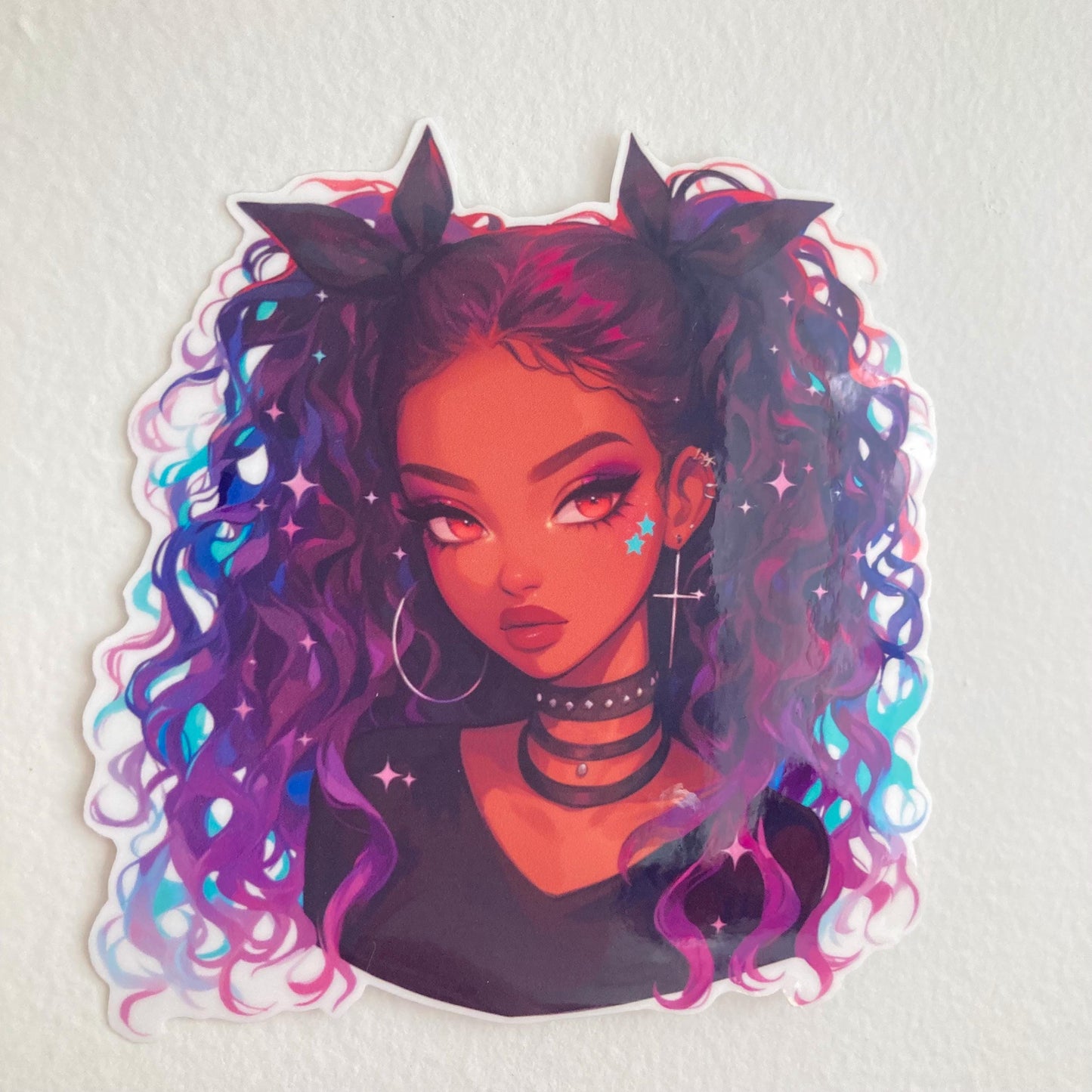 A sticker of a girl with curly purple and blue hair, chokers, hoop earrings, and star face decals, placed on a plain light-colored surface.