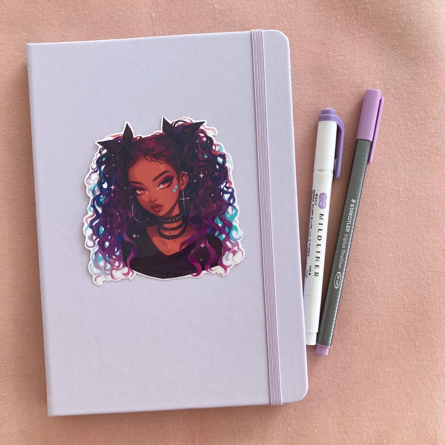 A sticker of a girl with curly purple and blue hair on a light teal notebook. Beside the notebook are a green marker and a gray pen, all set on a soft pink fabric background.