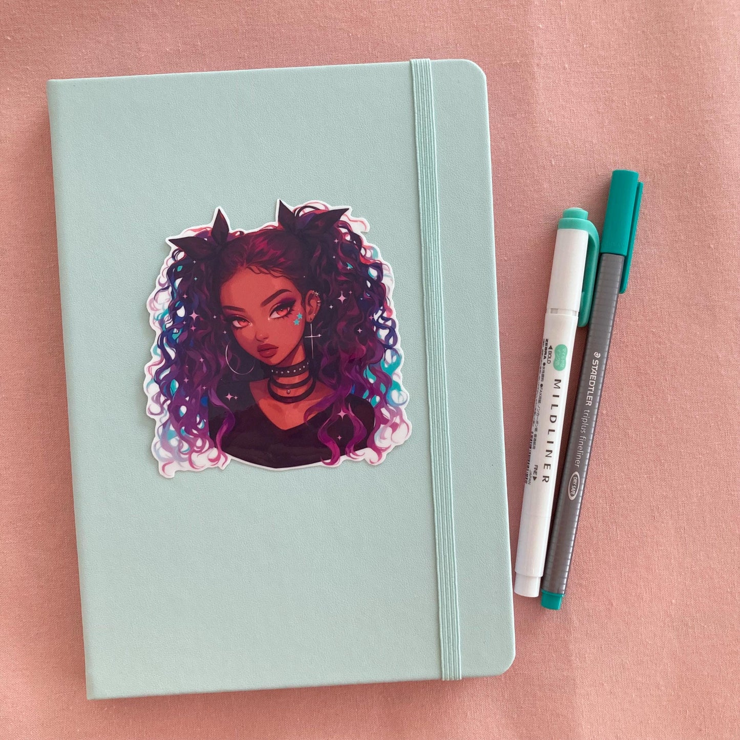 A sticker of a girl with curly purple and blue hair on a light teal notebook. Beside the notebook are a green marker and a gray pen, all set on a soft pink fabric background.