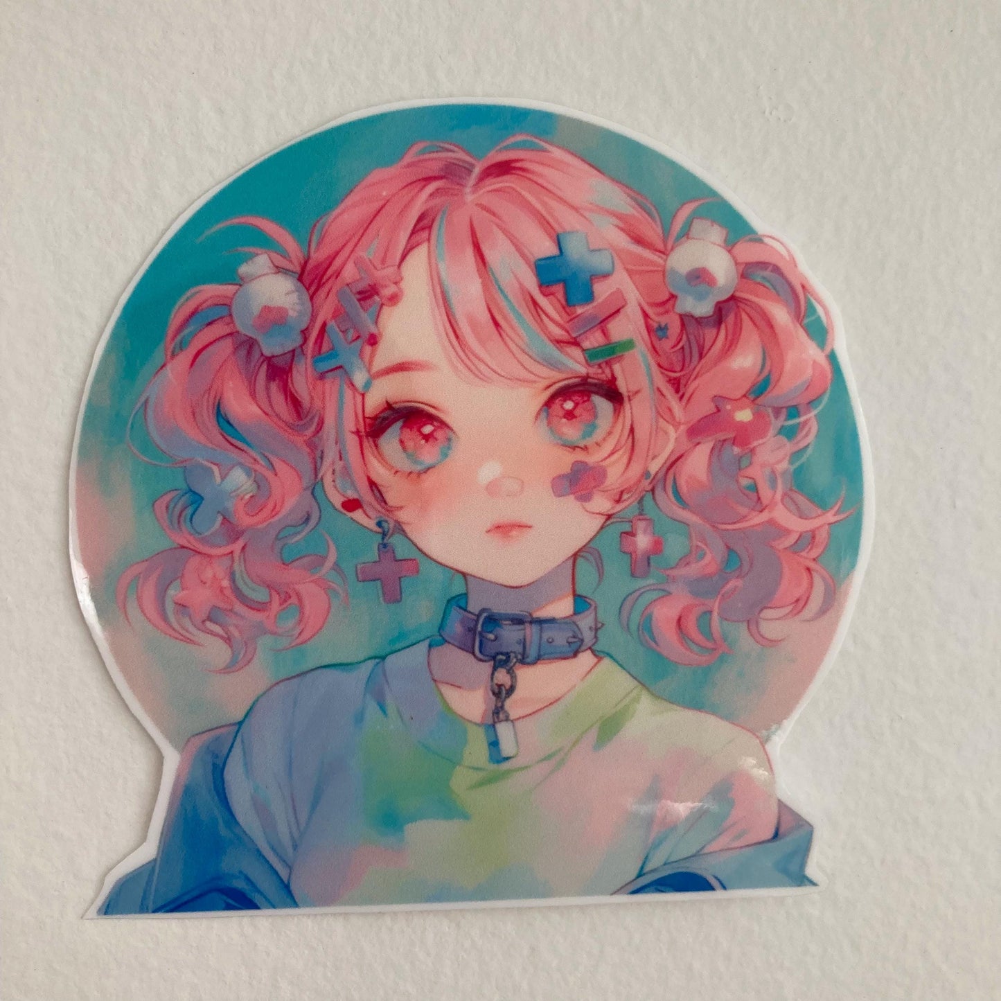 A sticker of a girl with pink pigtails, cross-shaped hair accessories, and a collar, wearing pastel-colored clothing. Placed against a plain light-colored background.