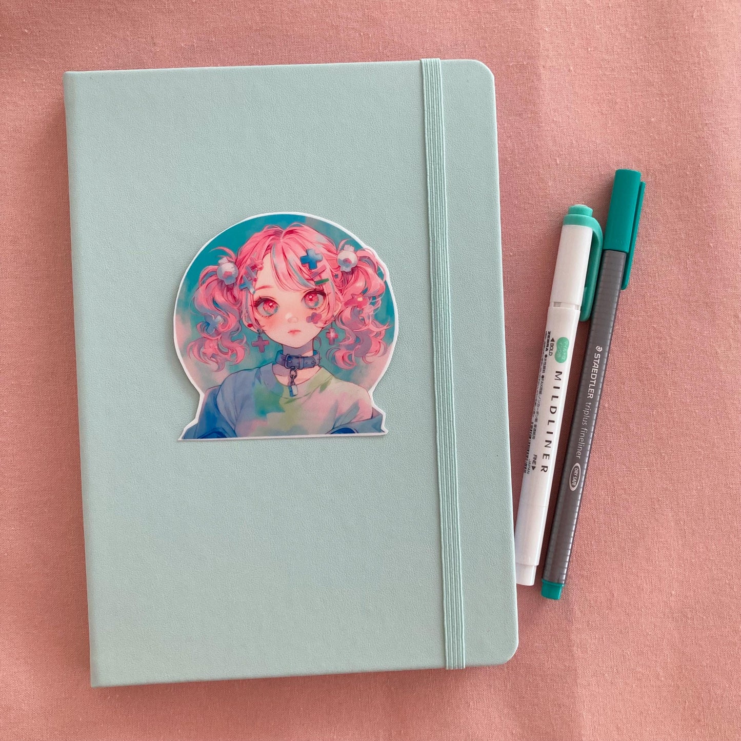 A sticker of a girl with pink pigtails and pastel clothing is placed on a light teal notebook. Beside it are a green marker and a gray pen, all on a soft pink fabric background.