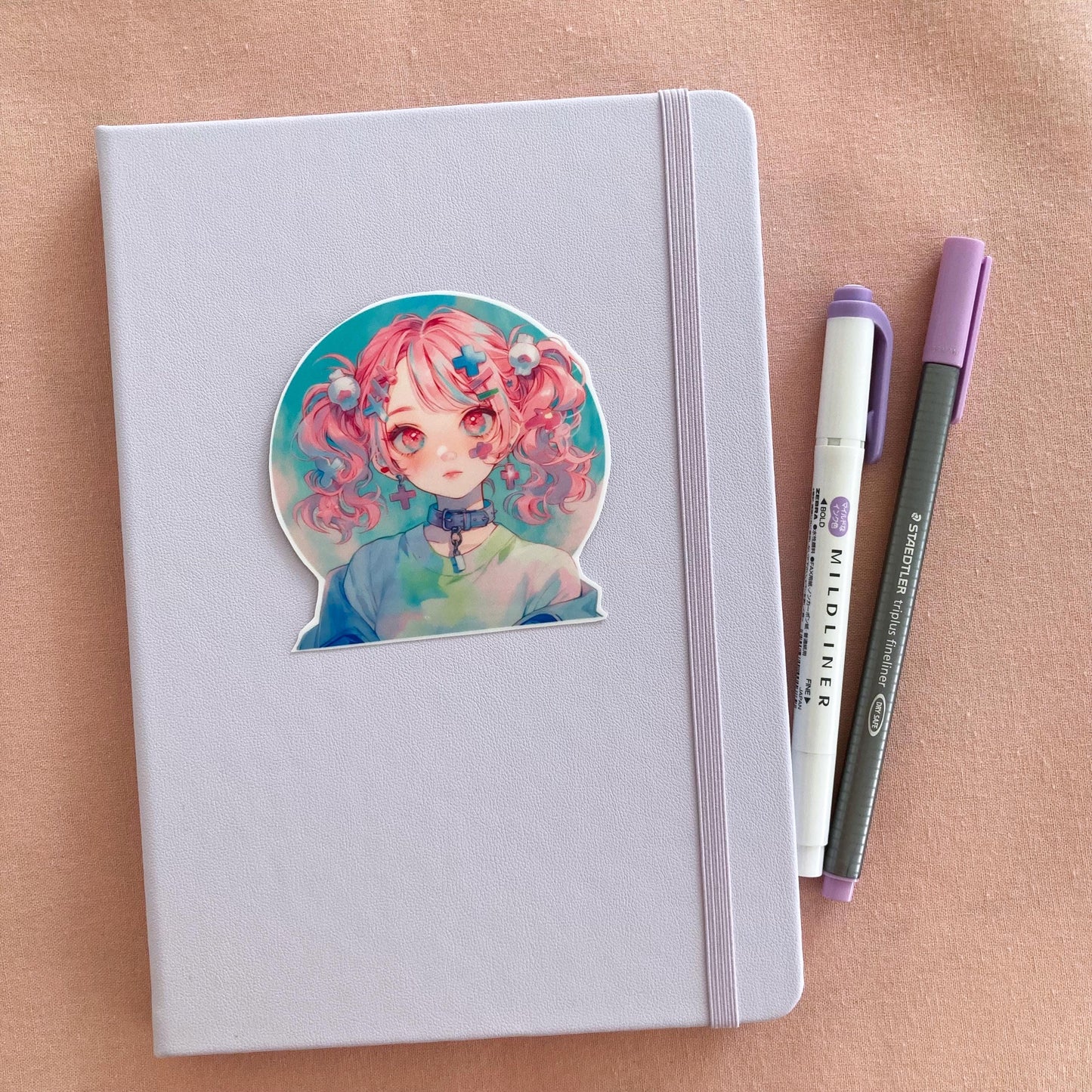 A sticker of a girl with pink pigtails and pastel clothes is placed on a light lavender notebook. Beside it are a purple marker and a gray pen, all set on a soft pink fabric background.