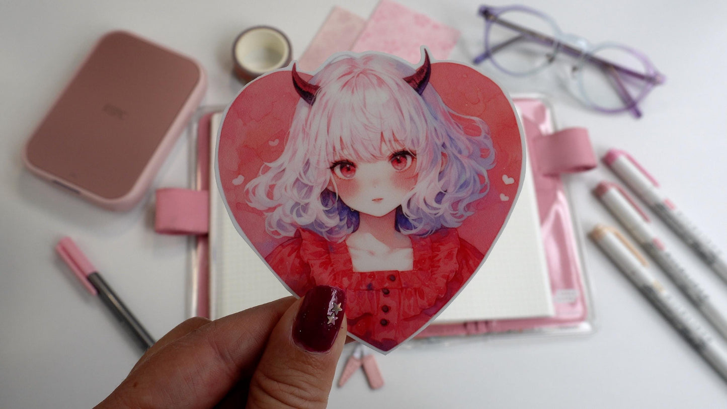 A hand holds a heart-shaped sticker of a girl with white hair, small red horns, and a red dress. The background includes a pink notebook, markers, washi tape, pink paper, a compact printer, and glasses. Nails are dark pink with a silver star accent.
