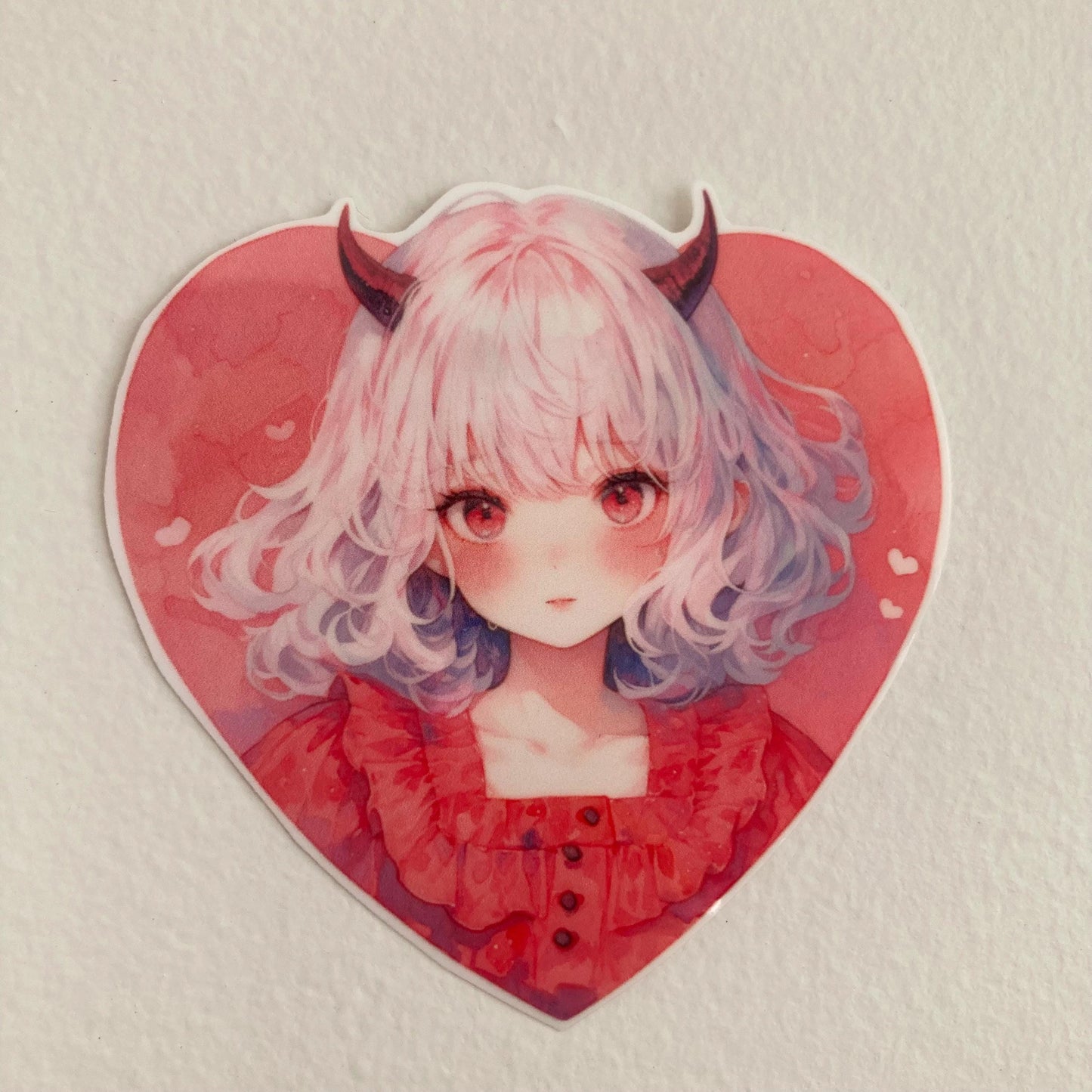 A heart-shaped sticker of a girl with white hair, small red horns, and a red dress, placed against a plain light-colored background.