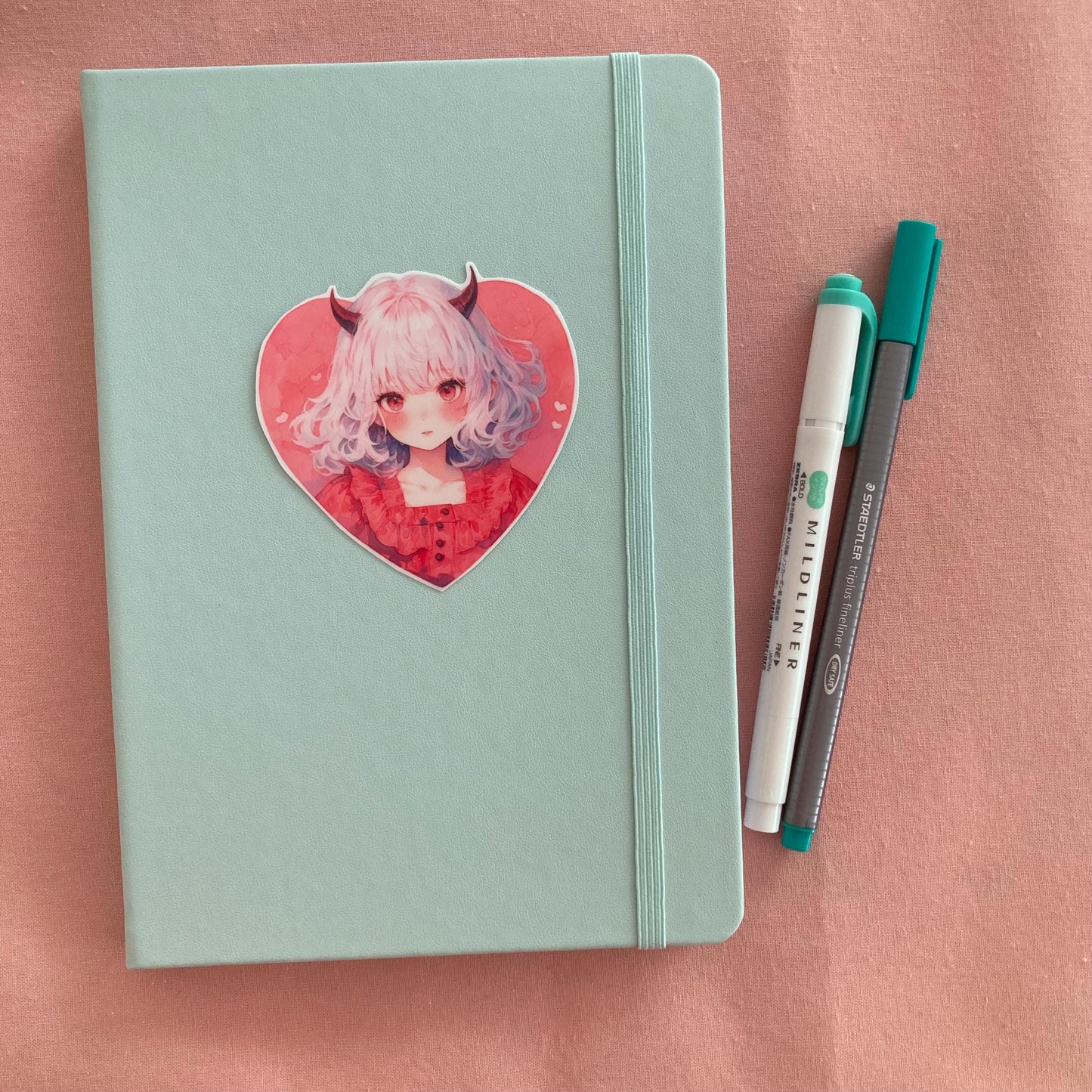 A heart-shaped sticker of a girl with white hair, red horns, and a red dress is placed on a light teal notebook. Beside the notebook are a green marker and a gray pen, all on a soft pink fabric background.