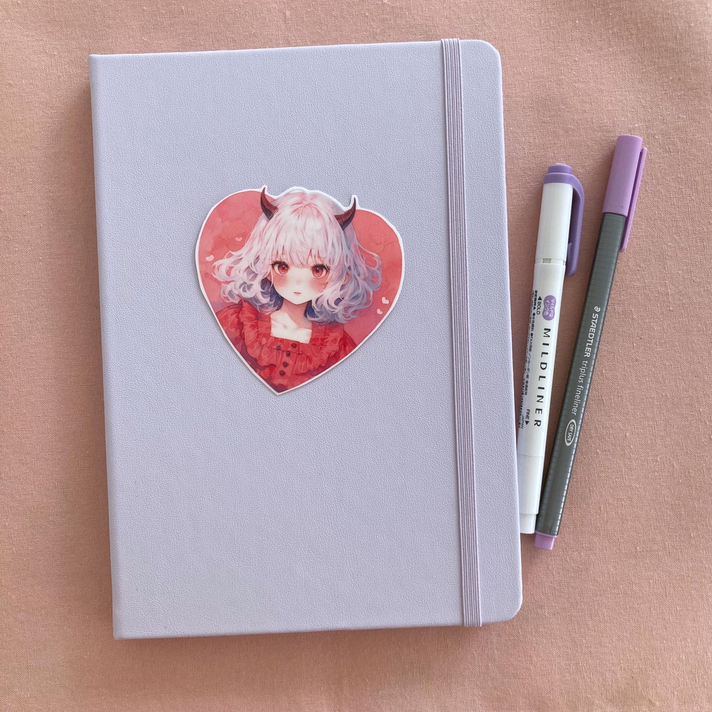 A heart-shaped sticker of a girl with white hair, red horns, and a red dress is placed on a light lavender notebook. Beside it are a purple marker and a gray pen, all on a soft pink fabric background.