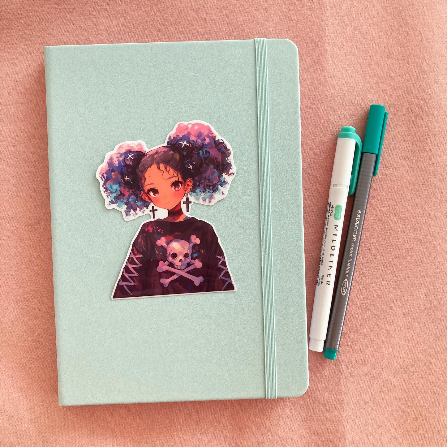 A pastel green notebook with a sticker of an anime-style character with purple and blue hair, cross earrings, and a skull-themed shirt, next to a green marker and gray pen on a pink fabric background.