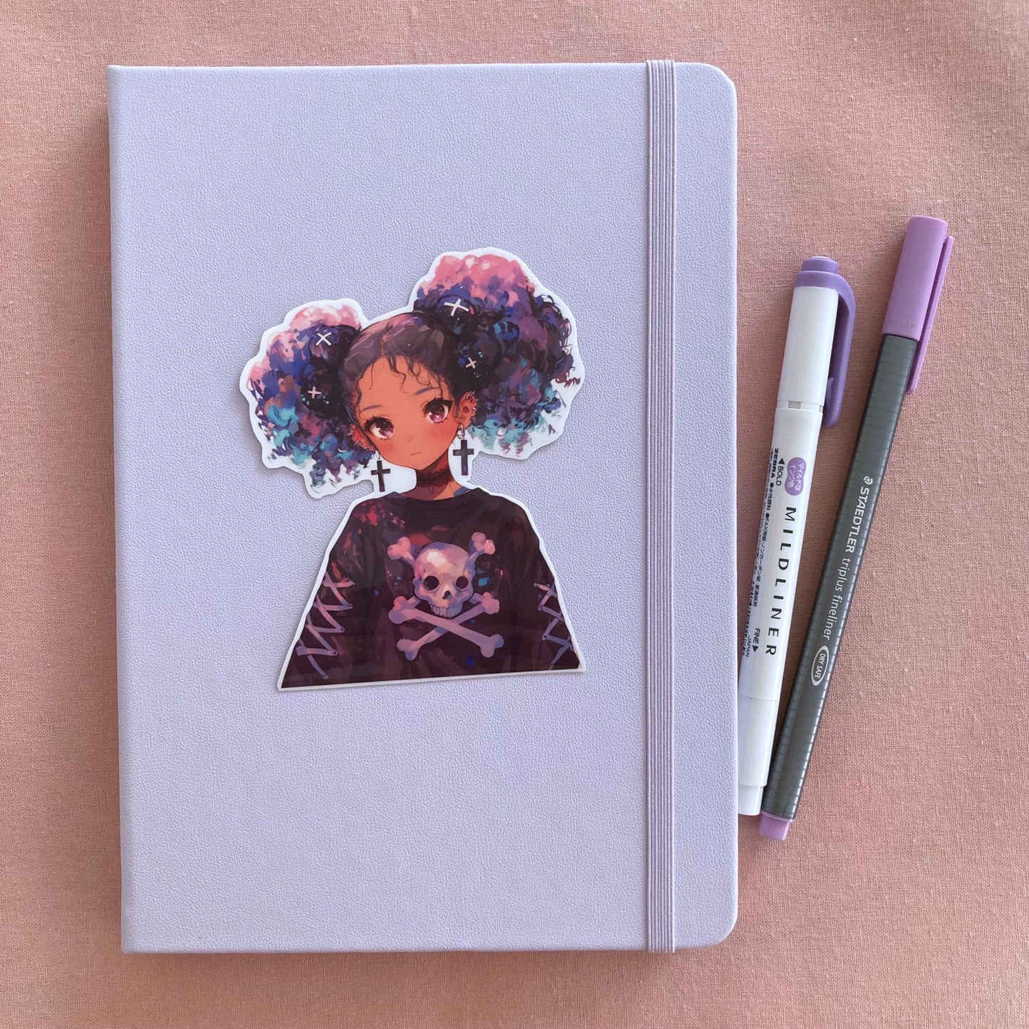 A pastel lavender notebook with a sticker of an anime-style character with purple and blue hair, cross earrings, and a skull-themed shirt, next to a purple marker and gray pen on a pink fabric background.