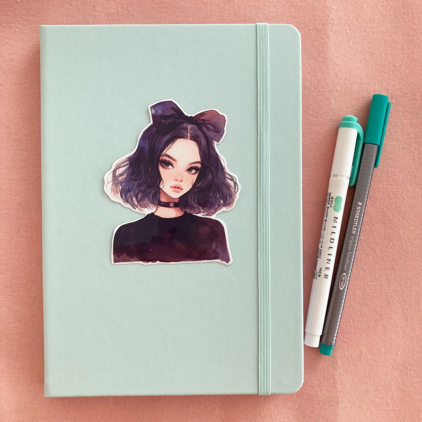 A pastel green notebook with a sticker of an anime-style character with dark purple hair, a large bow, and a black choker, next to a green marker and gray pen on a pink fabric background.
