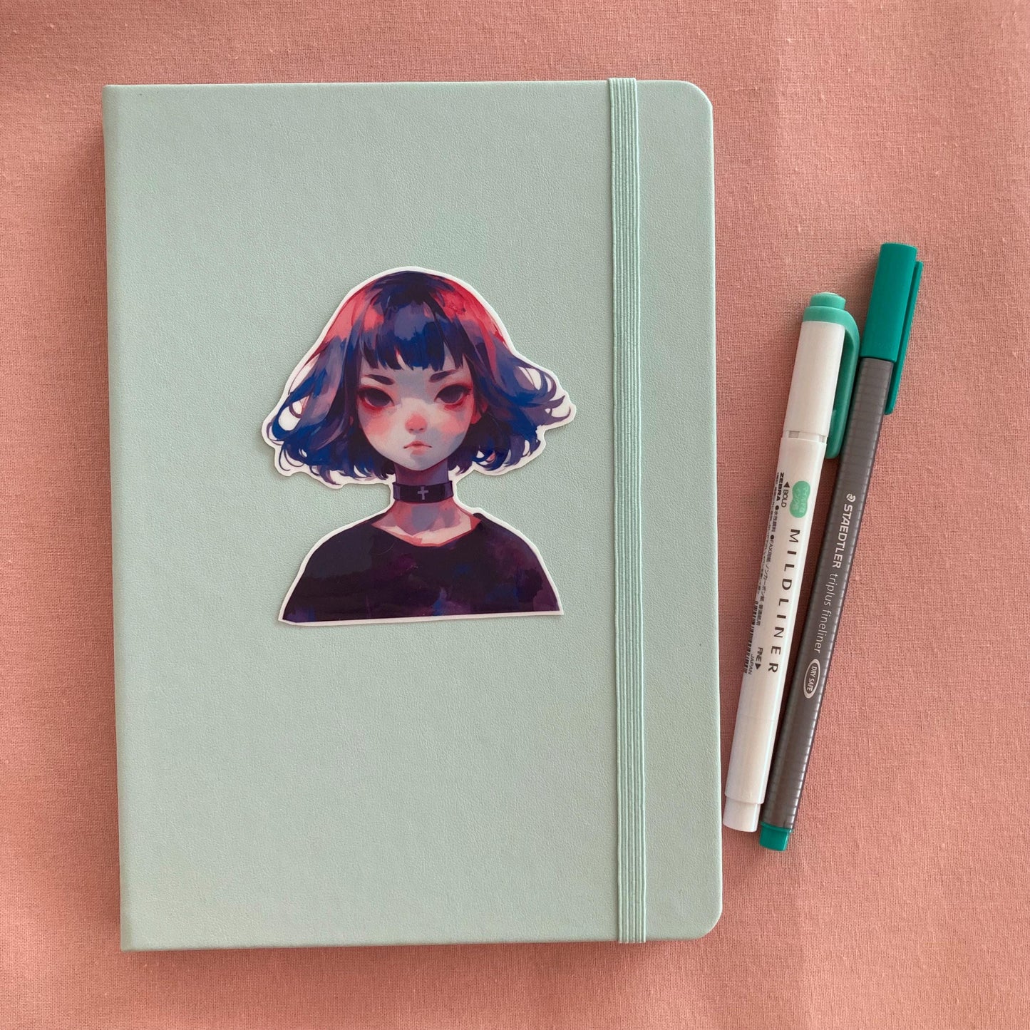 Sticker of an anime-style character with blue, pink, and purple hair and a black choker with a cross, placed on a mint green notebook. Two pens, one green and one gray, are to the right. The background is a soft pink surface.