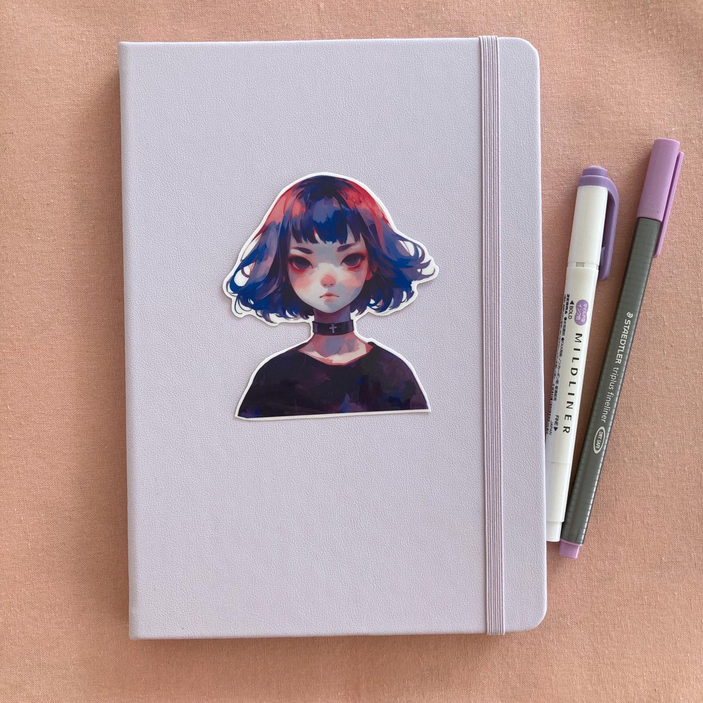 Sticker of an anime-style character with blue and pink hair and a black choker with a cross on a light lavender notebook. Two pens, one purple and one gray, are on the right side. Background is a soft pink surface.