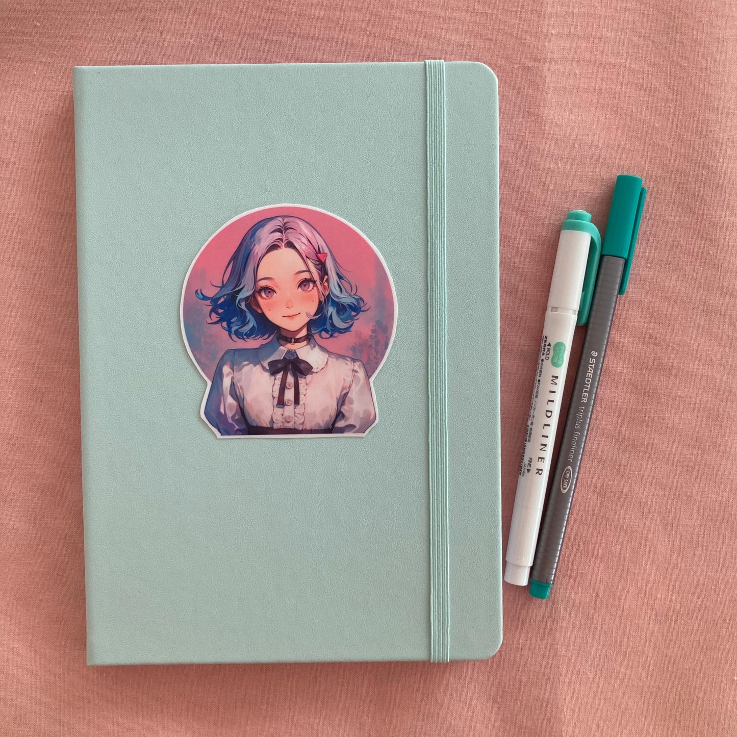 Sticker of an anime-style character with blue and pink hair, wearing a white blouse with a black ribbon on a light green notebook. Two pens, one green and one gray, are on the right side. The background is a soft pink surface.