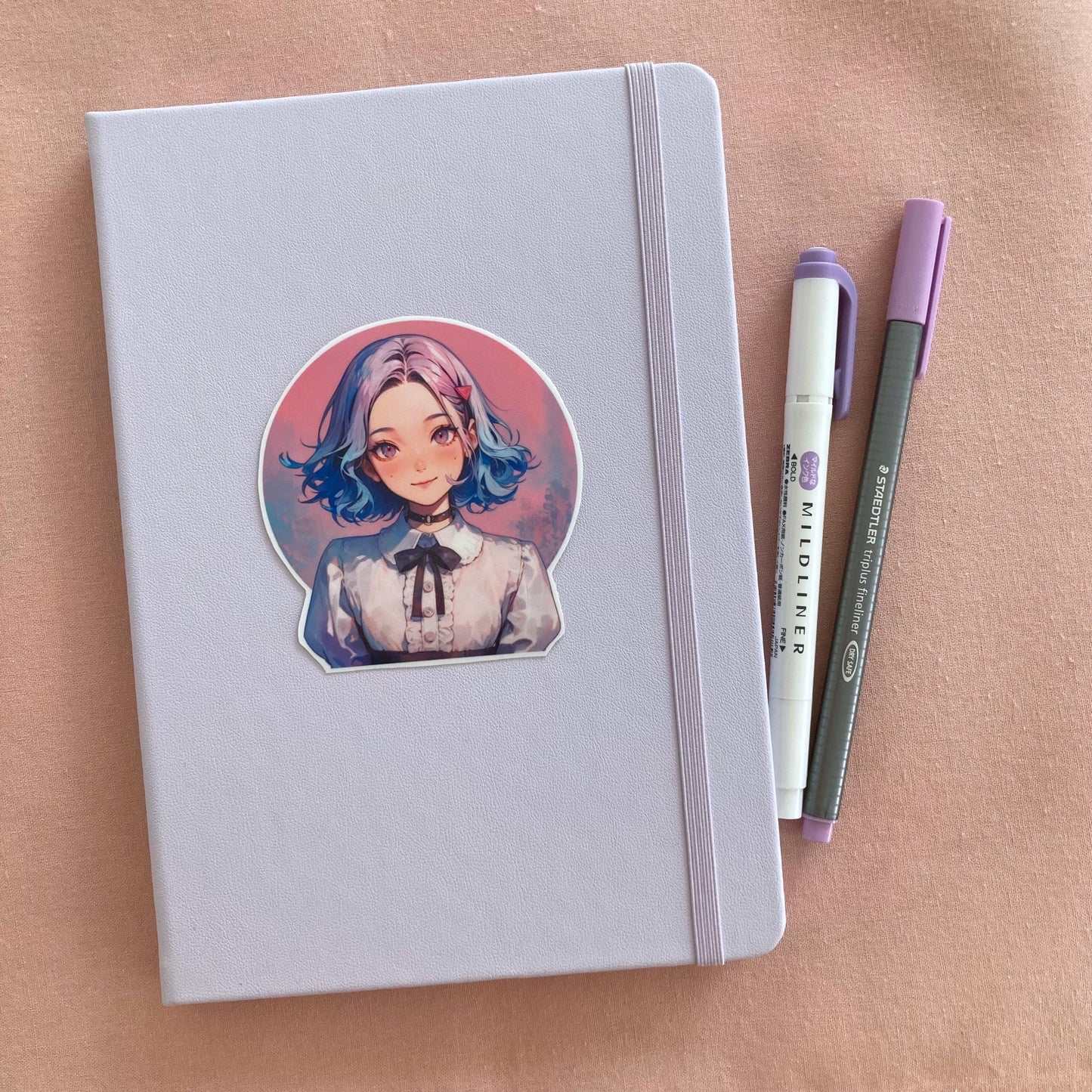 Sticker of an anime-style character with short blue and pink hair and wearing a white blouse with a black ribbon on a light lavender notebook. Two pens, one purple and one gray, are on the right side. Background is a soft pink surface.