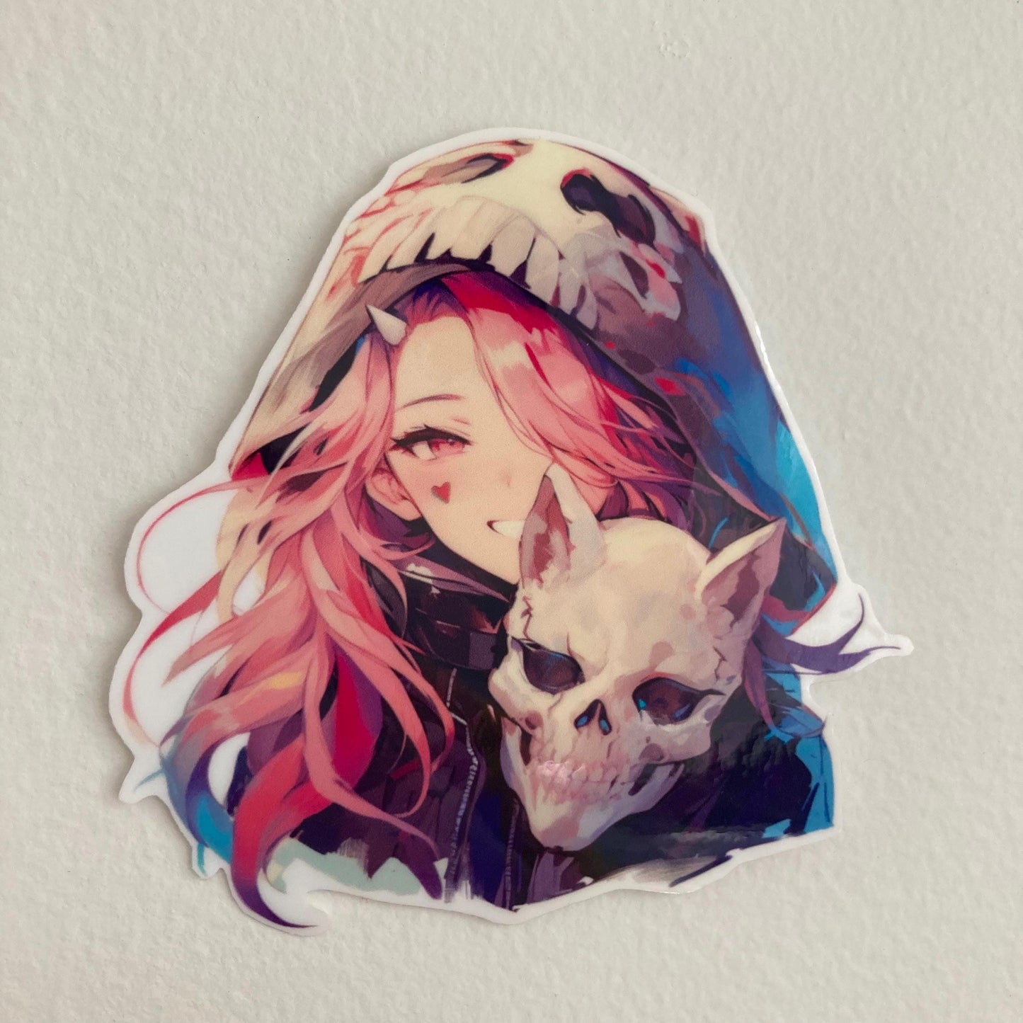 Sticker of an anime character with pink hair, wearing a skull-themed hood, holding a skull, and smiling with a heart mark on the cheek. Set against a plain light background.