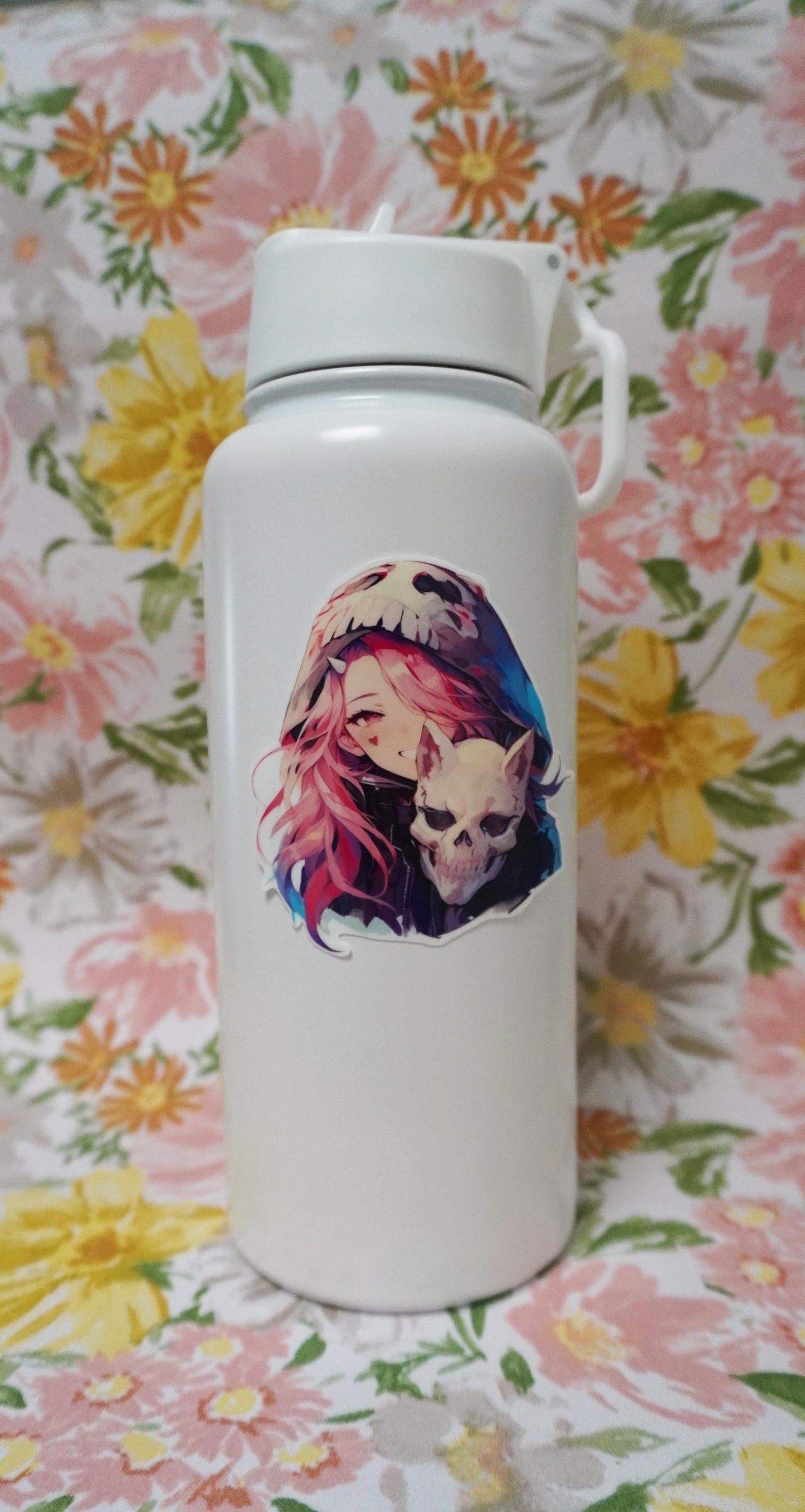 White bottle with a sticker of an anime character with pink hair, a skull-themed hood, and holding a skull. The background features a floral fabric with large pink, yellow, and green flowers.