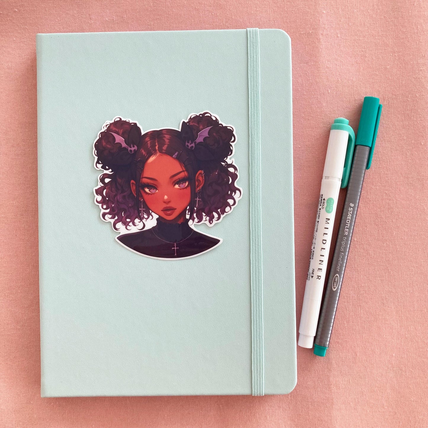 A mint green notebook with a sticker of a girl with curly buns, bat-shaped hair clips, and cross earrings on the cover. Beside the notebook are a white and green marker and a gray fineliner pen, all placed on a soft pink surface.