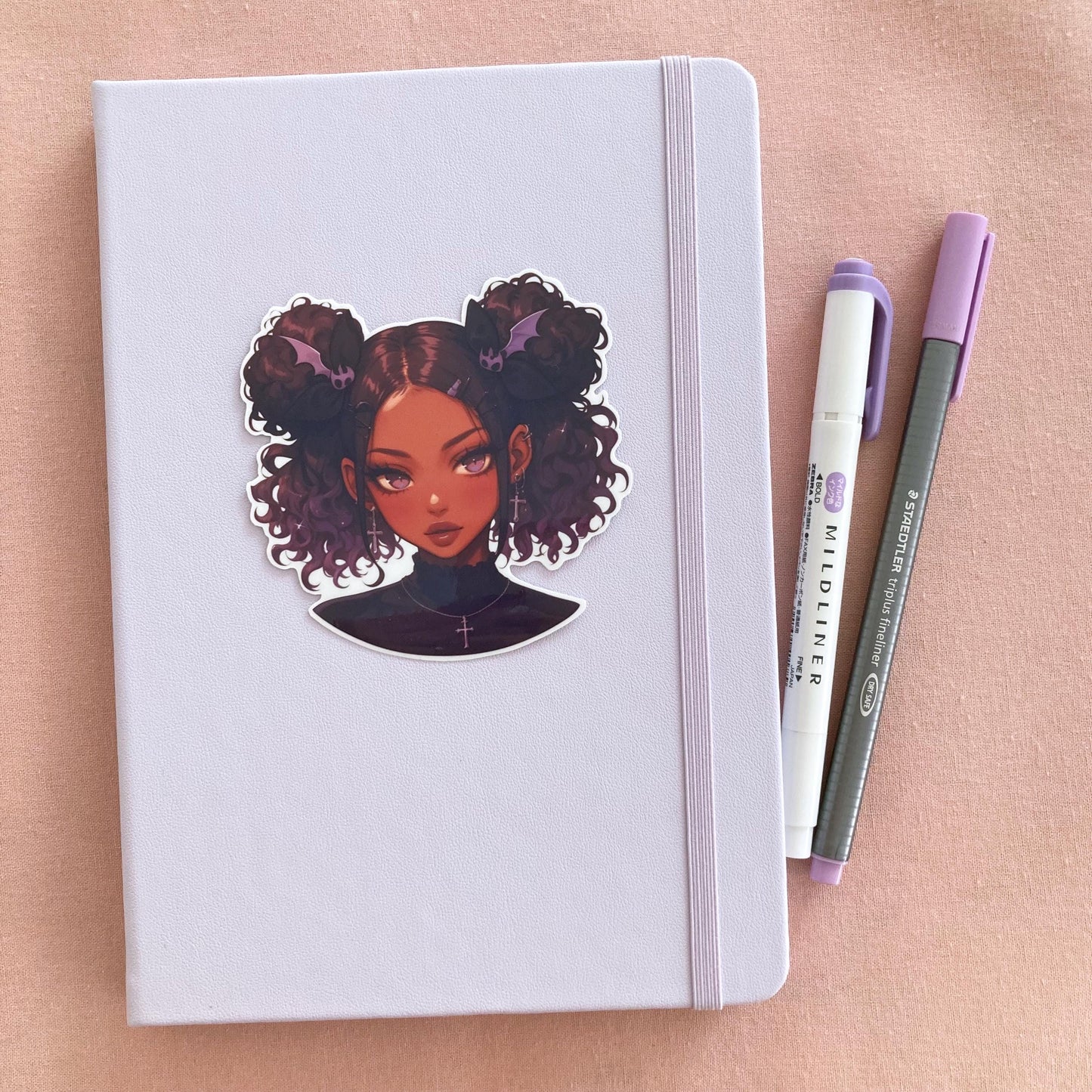 A light purple notebook with a sticker of a girl with curly buns, bat-shaped hair clips, and cross earrings on the cover. Next to the notebook are a white and purple marker and a gray fineliner pen, all on a soft pink surface.