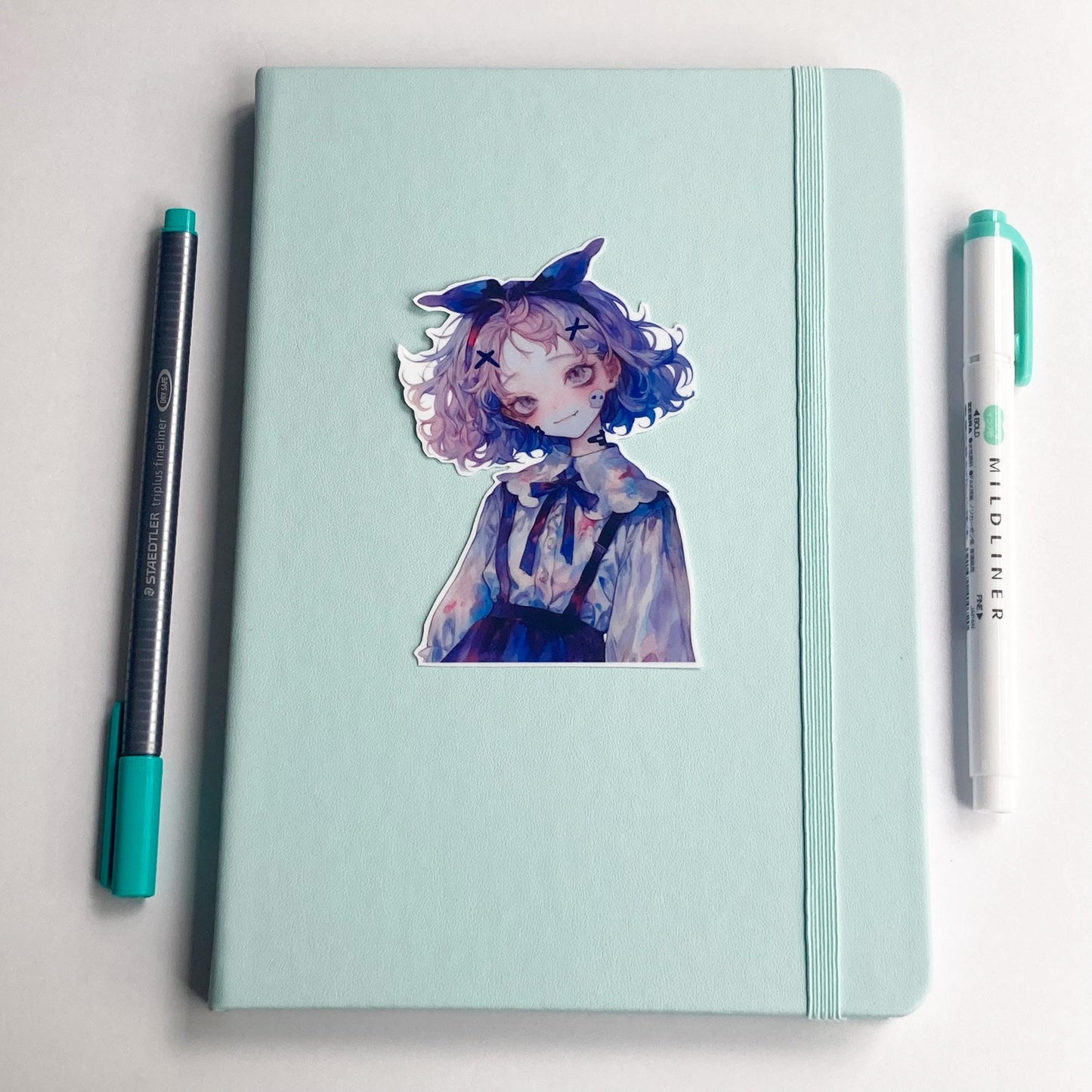 A sticker of an anime-style character is placed on a closed mint-green notebook with an elastic band. A gray pen with a teal cap is on the left side, and a white "Mildliner" marker with a teal cap is on the right side, both parallel to the notebook.