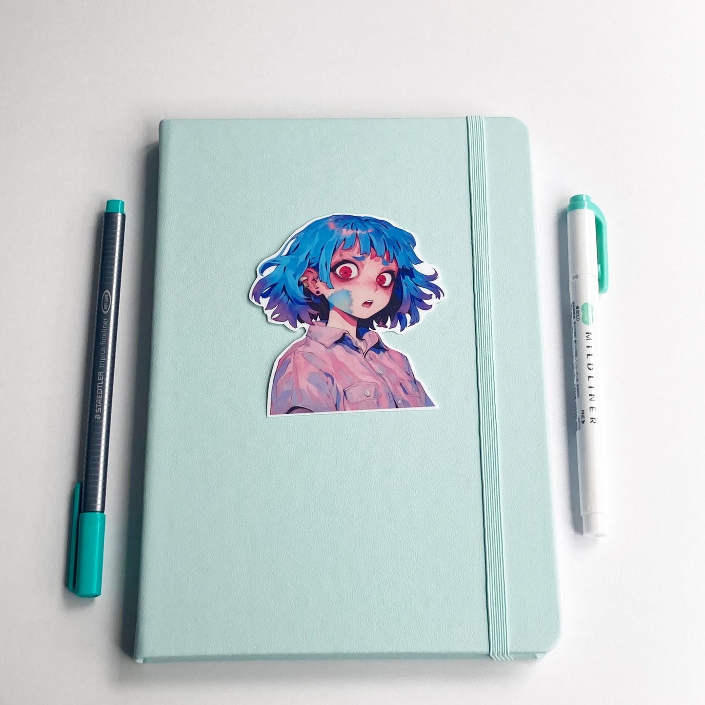 A sticker of an anime character with blue hair and red eyes is placed on a closed light teal notebook. A dark gray pen with a teal cap is on the left, and a teal marker is on the right. The background is a white surface.