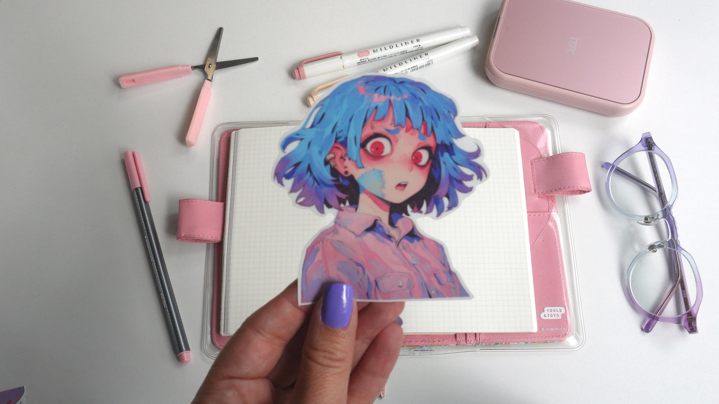 A hand with purple nails holds a colorful sticker of an anime character with blue hair and red eyes. The background includes a pink notebook, pastel pens, pink scissors, a small pink case, and purple-rimmed glasses, all arranged on a white surface