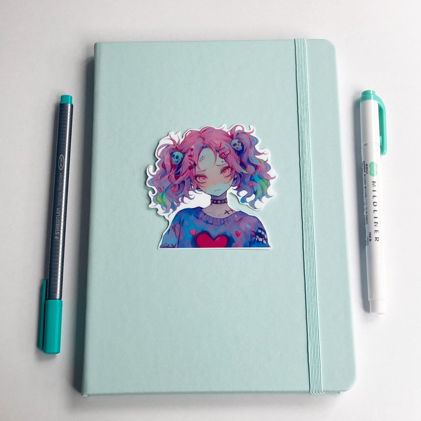 A sticker of an anime character with pink and multicolored hair and skull accessories is placed on a light teal notebook. A pen with a teal cap is on the left, and a teal marker is on the right.
