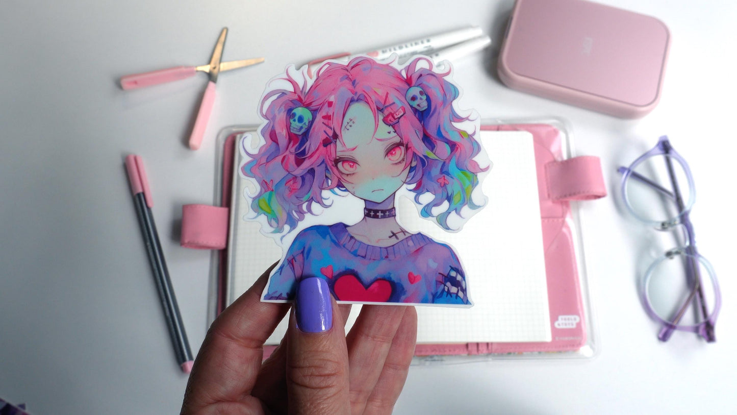 A hand with purple nails holds a sticker of an anime character with pink and multicolored hair, skull accessories, and a heart on their sweater. Background items include a pink notebook, pens, scissors, a case, and glasses on a white surface.